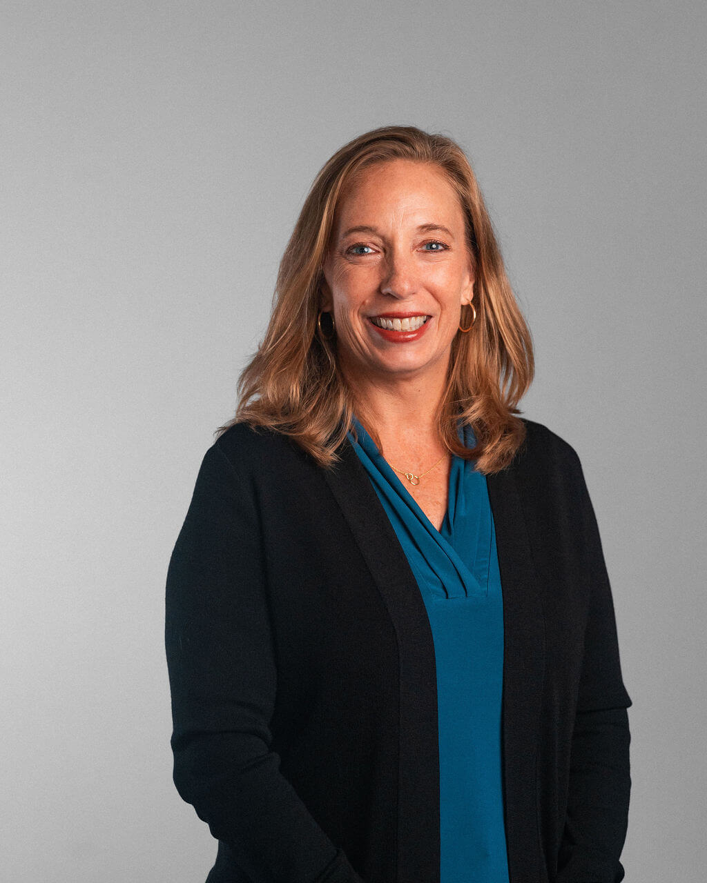 Wendy Paszkiewicz, a woman with long light brown hair, stands facing forward in a teal blouse and black cardigan, smiling against a plain light gray background.