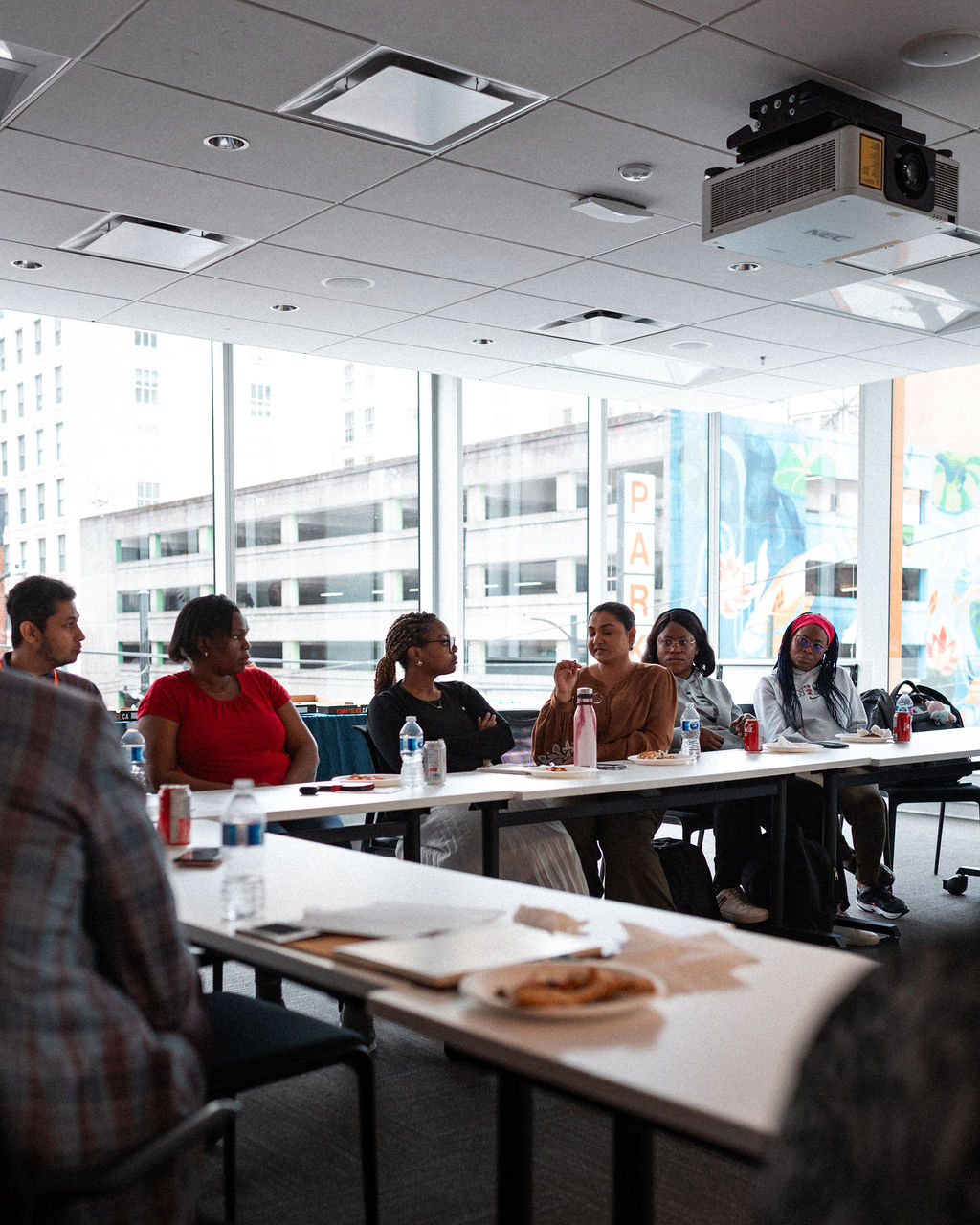 A group of people sit around tables in a modern conference room having a discussion; drinks and papers are on the tables. Large windows provide natural light.