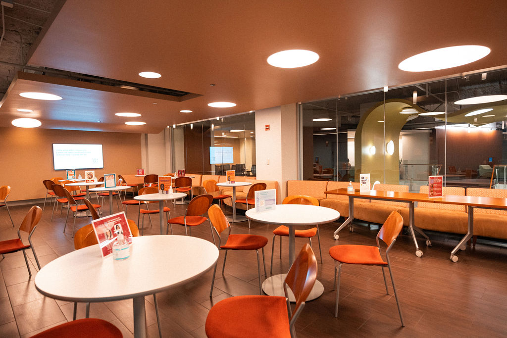 A modern lounge area with round white tables, orange chairs, and tan cushioned benches under circular ceiling lights. Informational signs are placed on the tables.