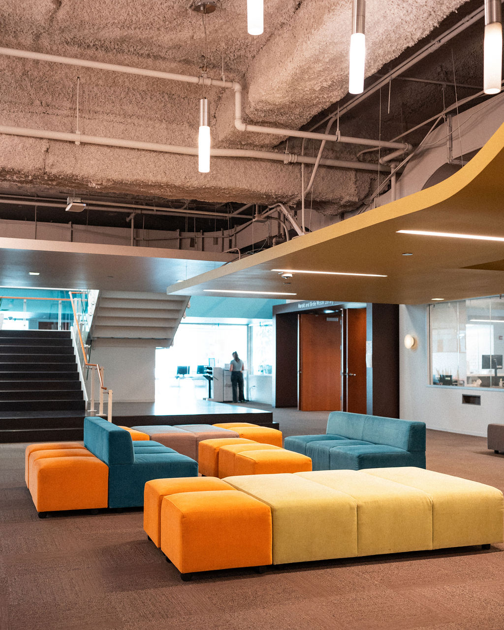 Modern office lounge with colorful modular seating, a staircase, ceiling lights, exposed pipes, and a person standing near glass doors in the background.