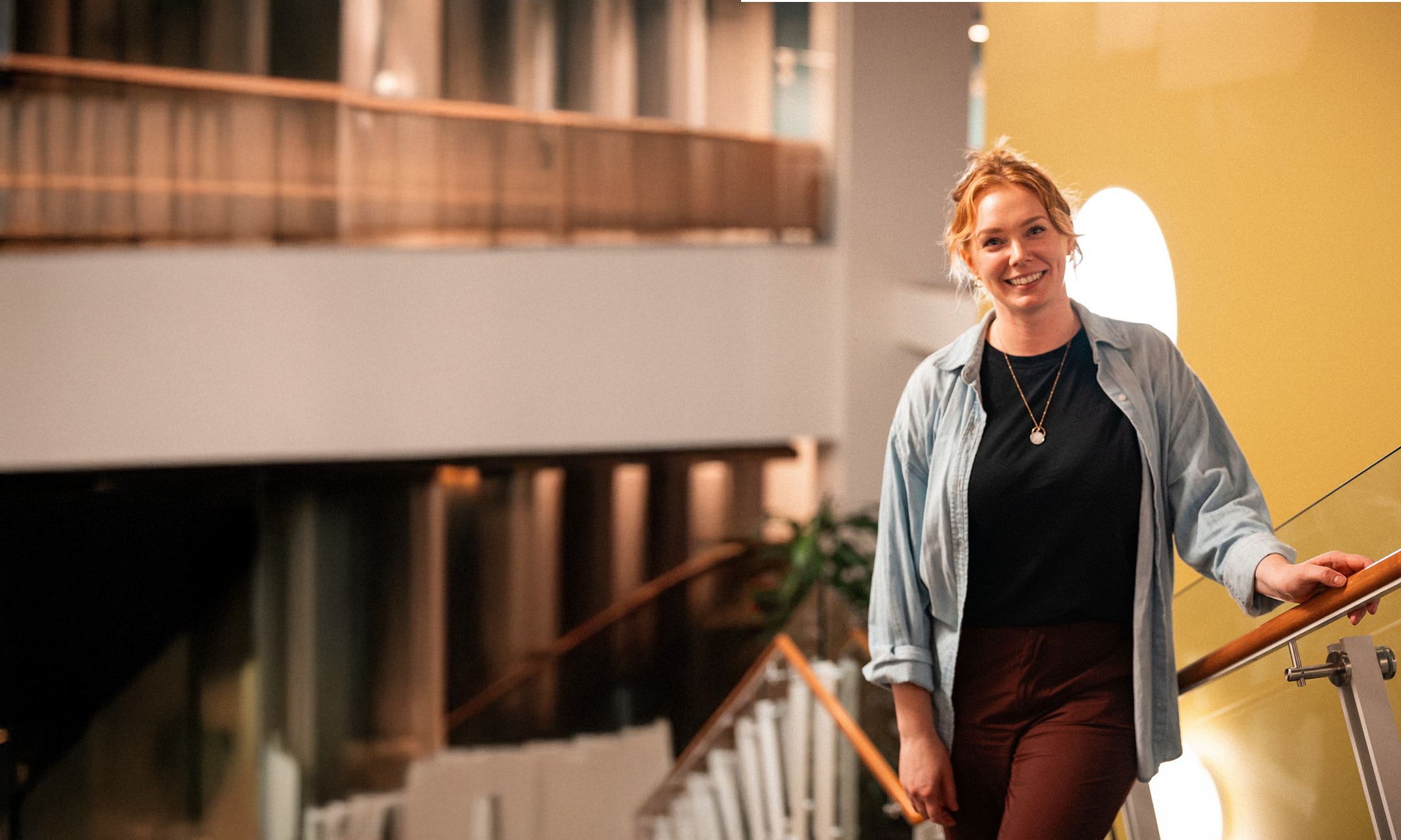 A person with light hair, wearing a blue shirt and dark pants, stands smiling on an indoor staircase with modern architecture in the background.