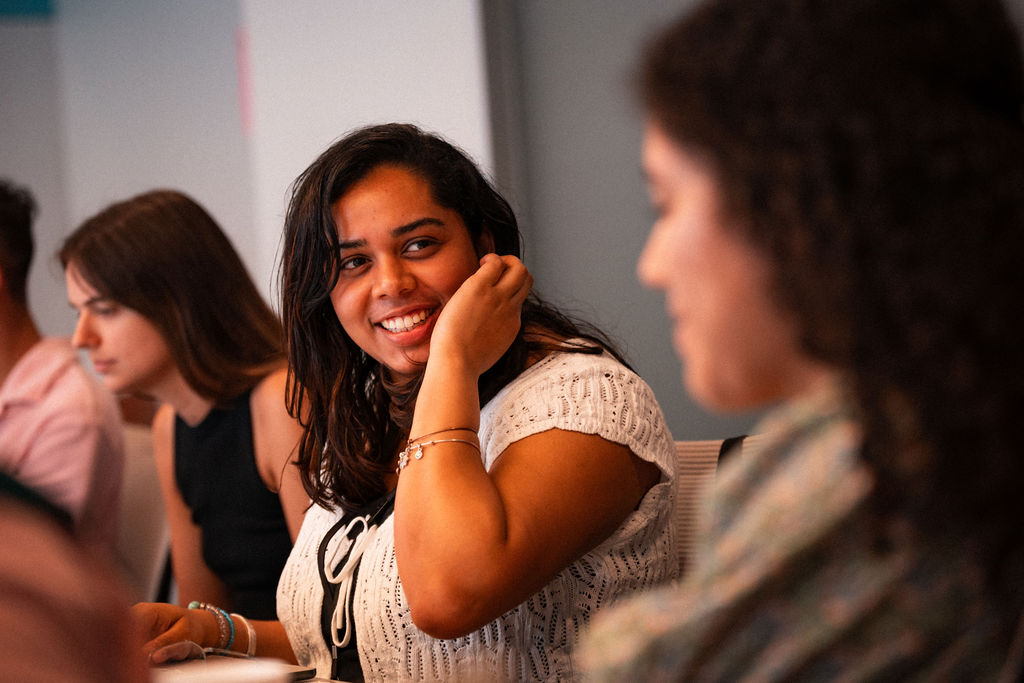 A woman in a white top smiles and looks at another person during a conversation about alumni relations in a group setting.