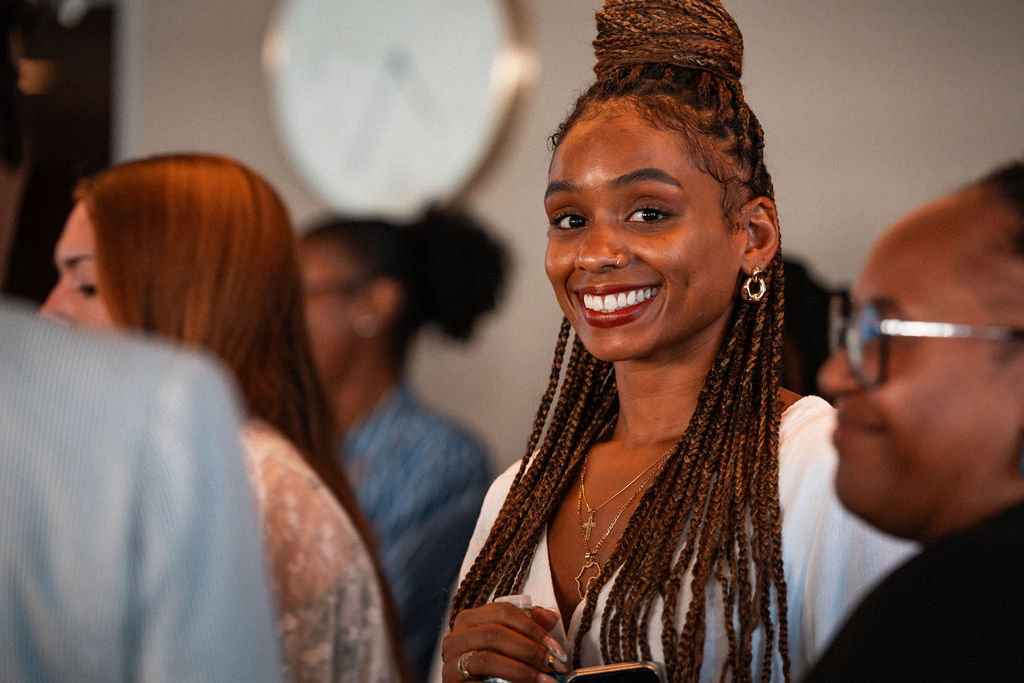 A woman with long braids smiles at the camera in a room with other people; a blurred clock is visible in the background.