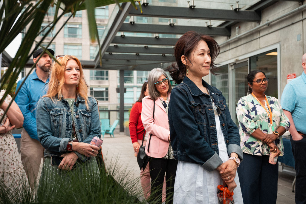 A group of adults, both men and women, stand outdoors on a rooftop, attentively listening to something out of frame that reflects shared values. Some hold phones and wear casual clothing.