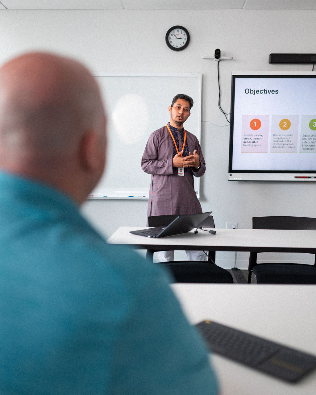 A man stands near a screen displaying objectives on institutional advancement and presents to a seated person in a conference room with laptops and a whiteboard, focusing on alumni relations and fundraising strategies.