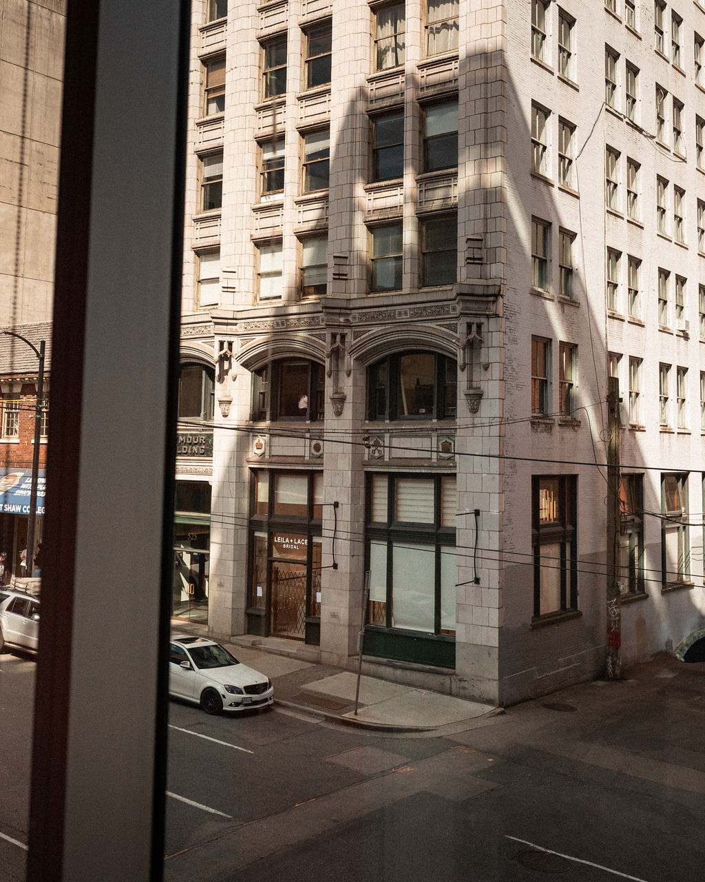 View from a window showing a street corner with a white multi-story building, parked cars, and storefronts on the ground level in daylight.