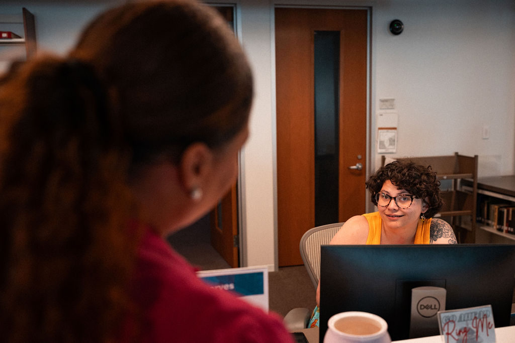 A person with curly hair and glasses sits at a desk, speaking to another person standing in front of them in an office setting.