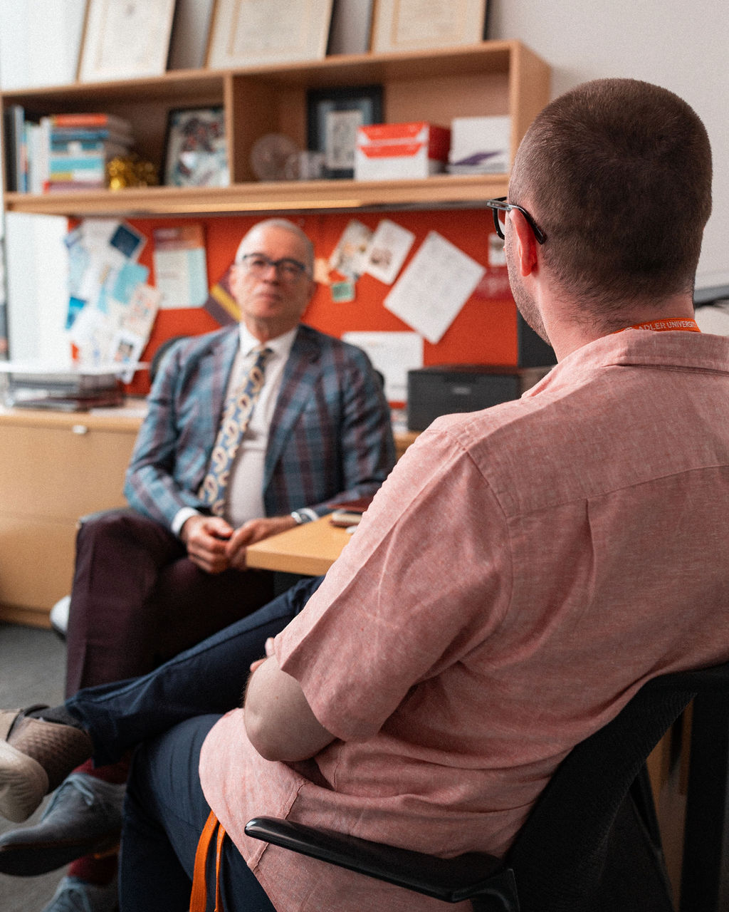 Two men sit facing each other in an office, one in a checked suit behind a desk and the other in a casual shirt, engaged in conversation.