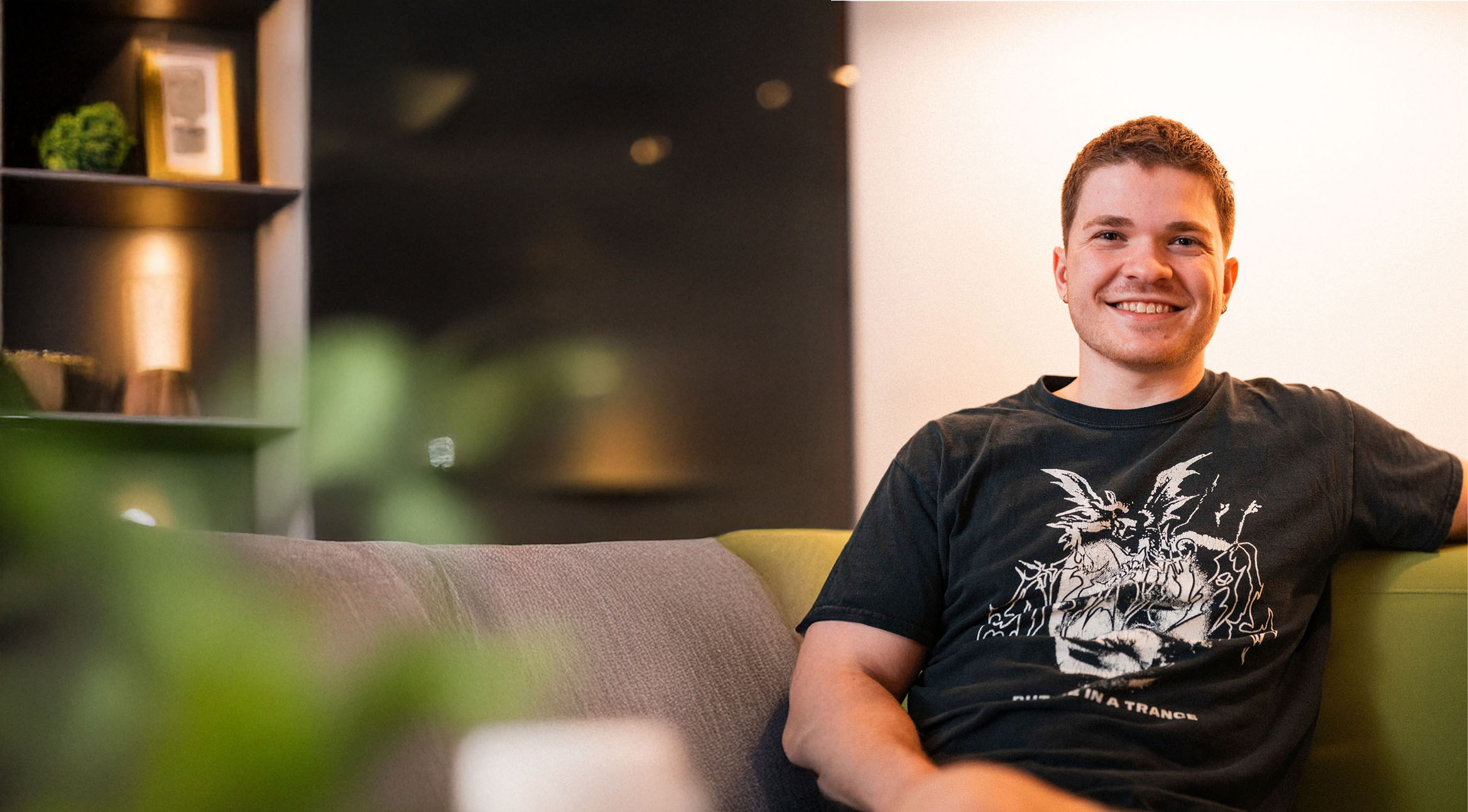 A man sits on a couch, smiling at the camera in his black graphic t-shirt. The background features shelves and soft lighting, reflecting a space where vision and values come together.