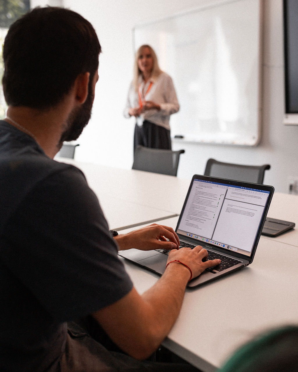 A person types on a laptop at a conference table while another person stands and speaks in front of a whiteboard.