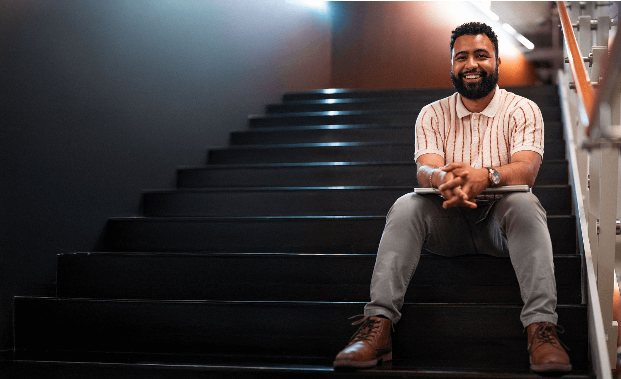 A man with a beard sits on a dark staircase, smiling at the camera, holding a laptop on his lap.