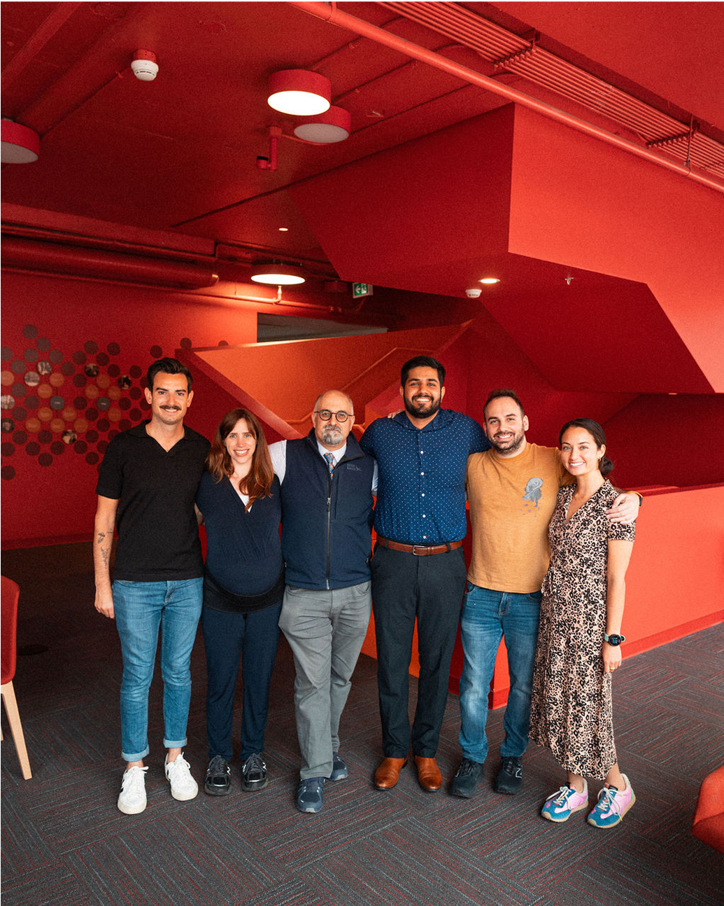 Six people stand side by side, smiling, in a brightly lit room with red walls and staircase in the background.