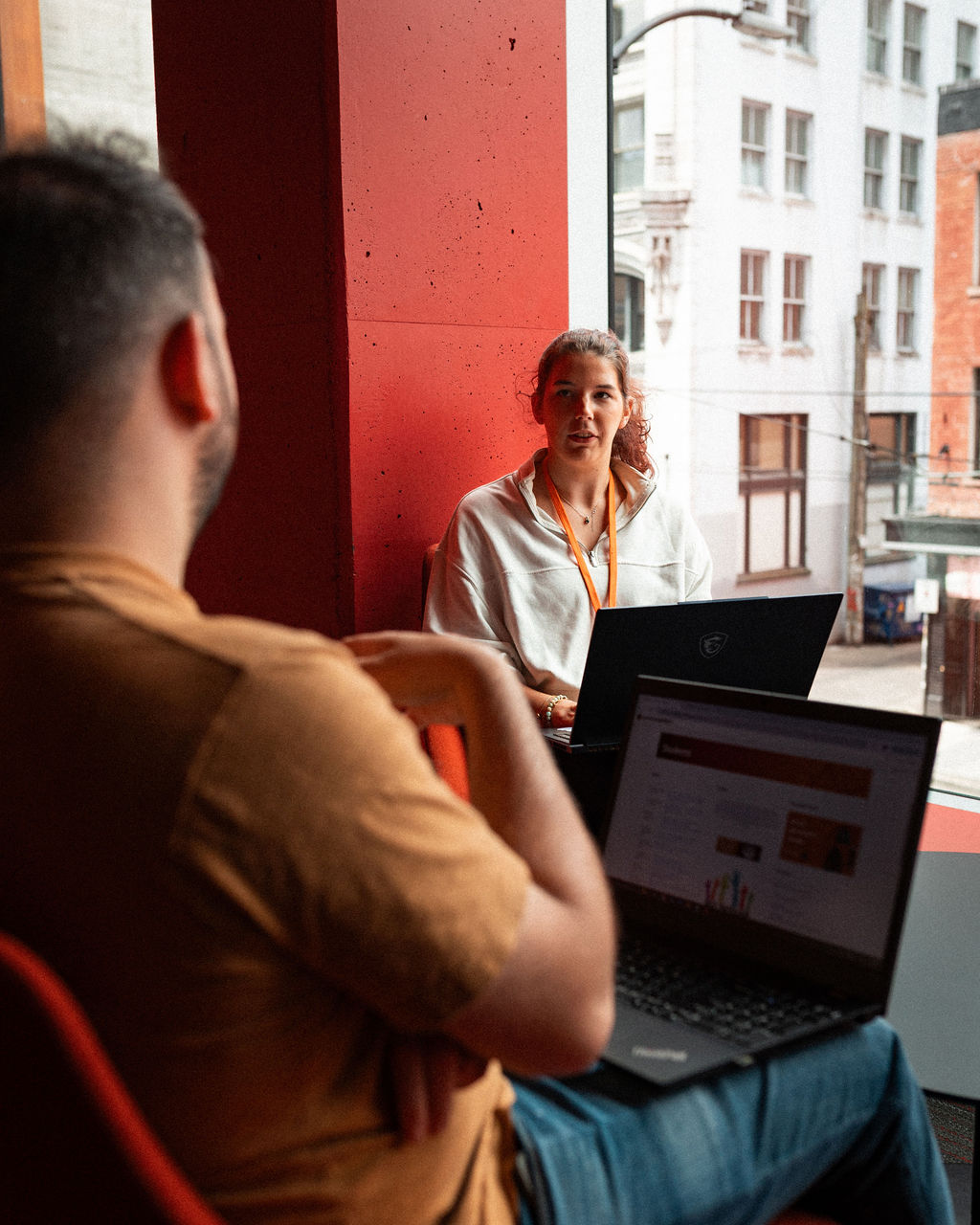 Two people sit by a window with laptops, engaged in conversation about overseas education. Urban buildings are visible outside through large windows.