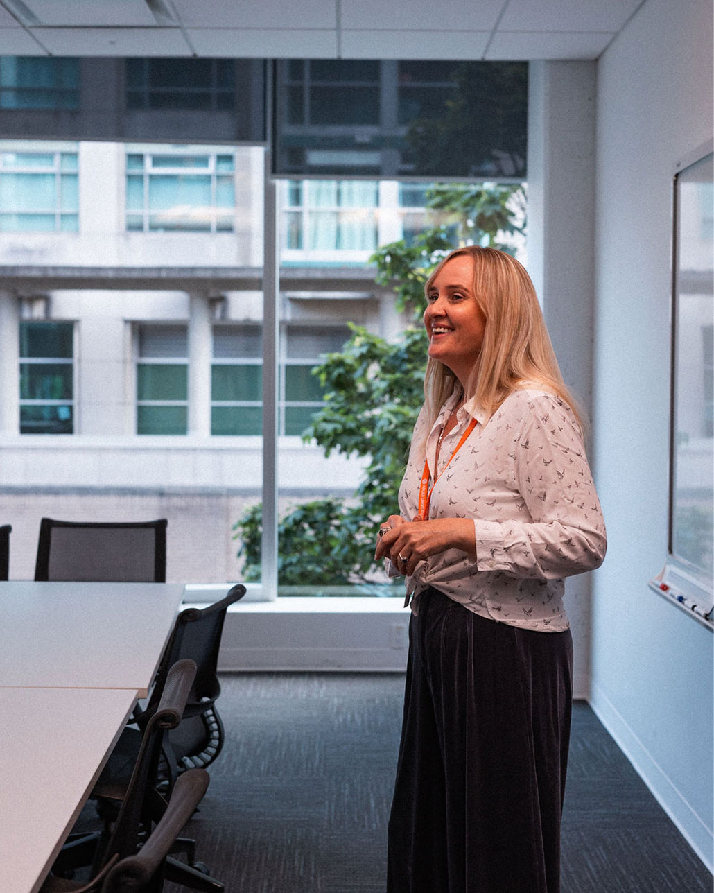A woman stands and smiles in a modern conference room next to a whiteboard, with large windows showing a building and trees outside.
