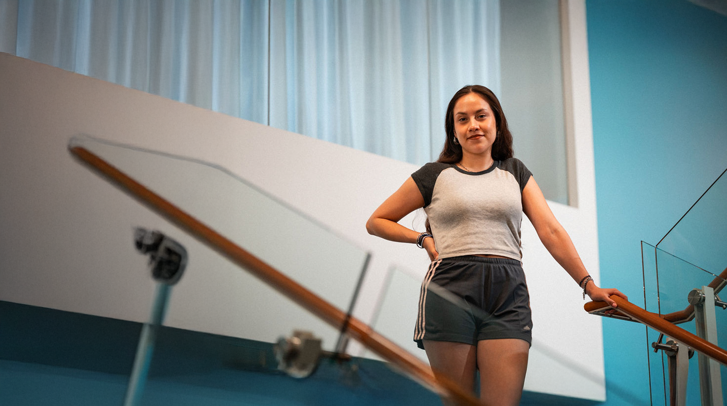 A young woman in athletic wear stands on an indoor staircase, holding the railing and looking at the camera. The background features blue walls and large windows with white curtains.