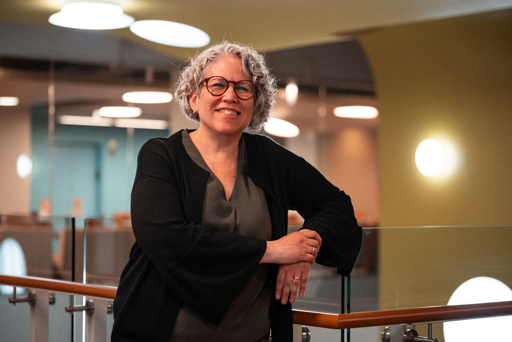 A woman with short curly gray hair and glasses, wearing a dark outfit, stands smiling by a glass railing in a modern indoor setting with round lights in the background.