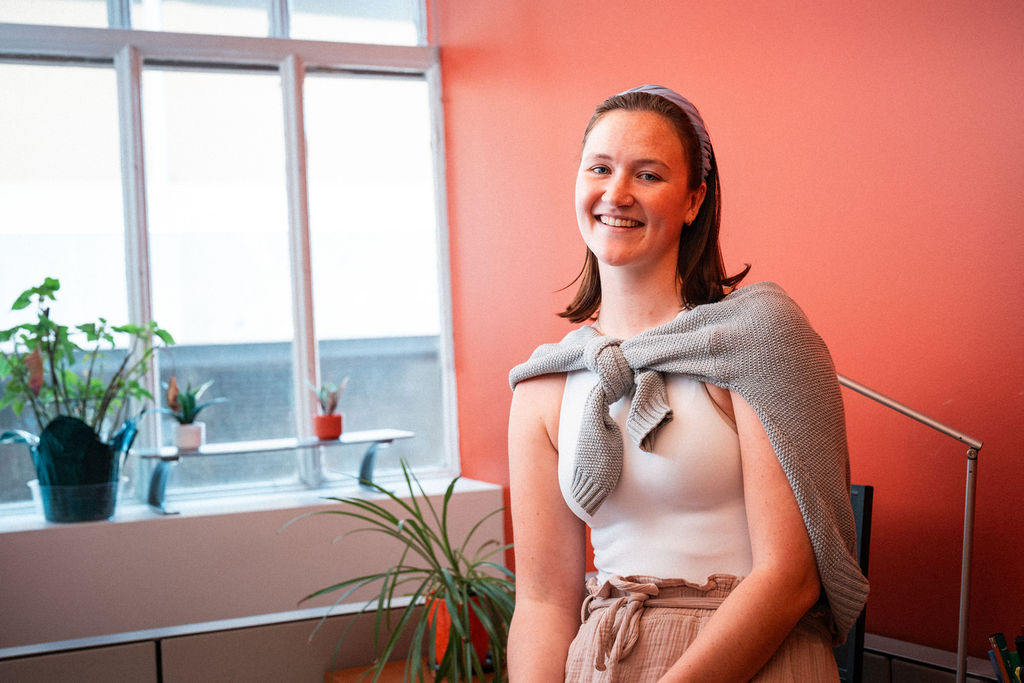 A woman wearing a headband and sweater draped over her shoulders sits and smiles in a room with a coral wall and several potted plants by a window.