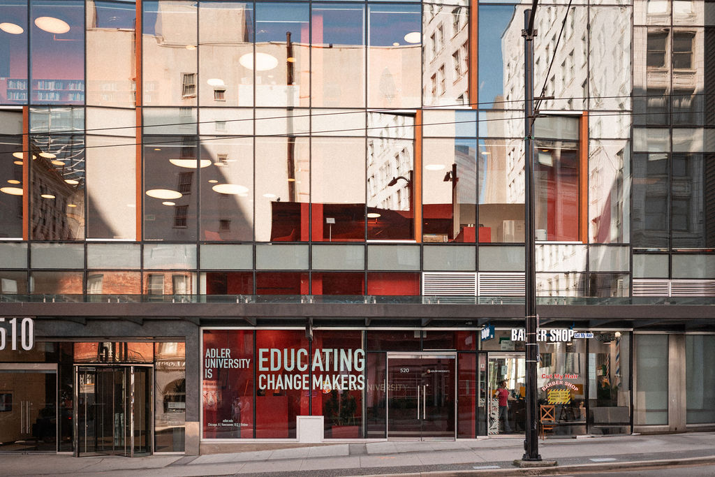 Street view of a modern glass building with Adler University signage reading "Educating Change Makers" and a barbershop entrance visible at street level.