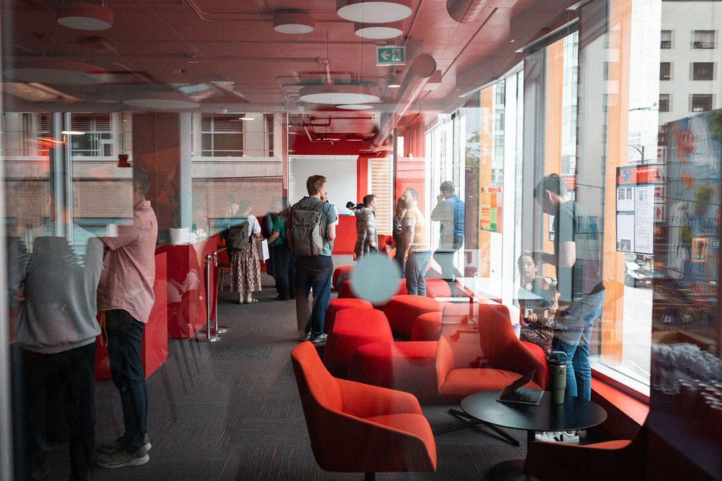 People stand and talk in a modern lounge with red chairs and large windows during the day. Some look at posters on the windows while others interact in small groups.