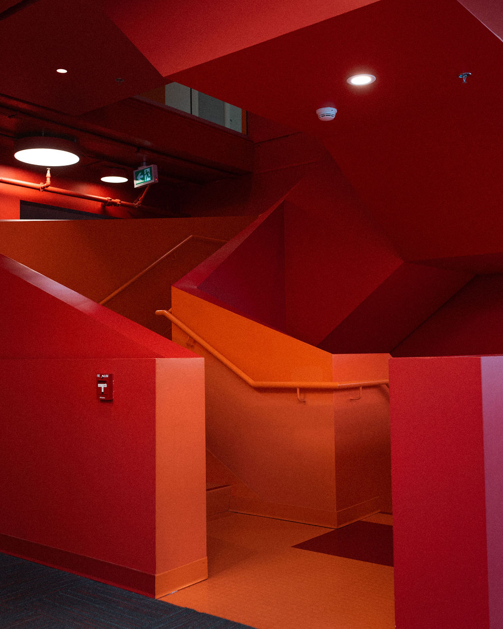 A stairwell with red and orange walls, ceiling, and railing, featuring modern geometric lines and illuminated by ceiling lights.