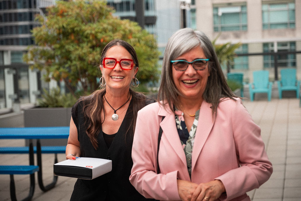 Two women stand and smile on an outdoor urban patio, embracing their shared vision. One holds a box; both wear glasses and casual clothing. Buildings are visible in the background.