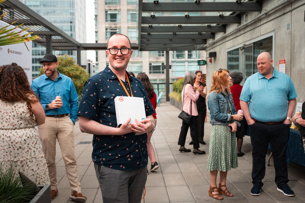 A man in glasses holds a white box and smiles at an outdoor event, where people are socializing in the background, celebrating shared mission and values.
