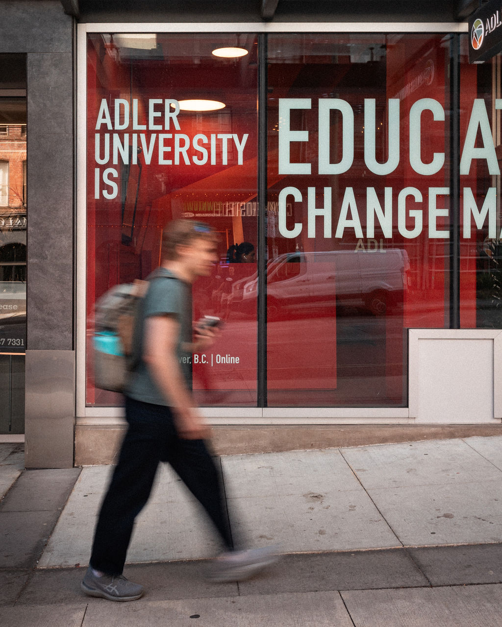 A person with a backpack walks on the sidewalk in front of Adler University, where large red and white text on the windows promotes education, change, and welcomes international students.