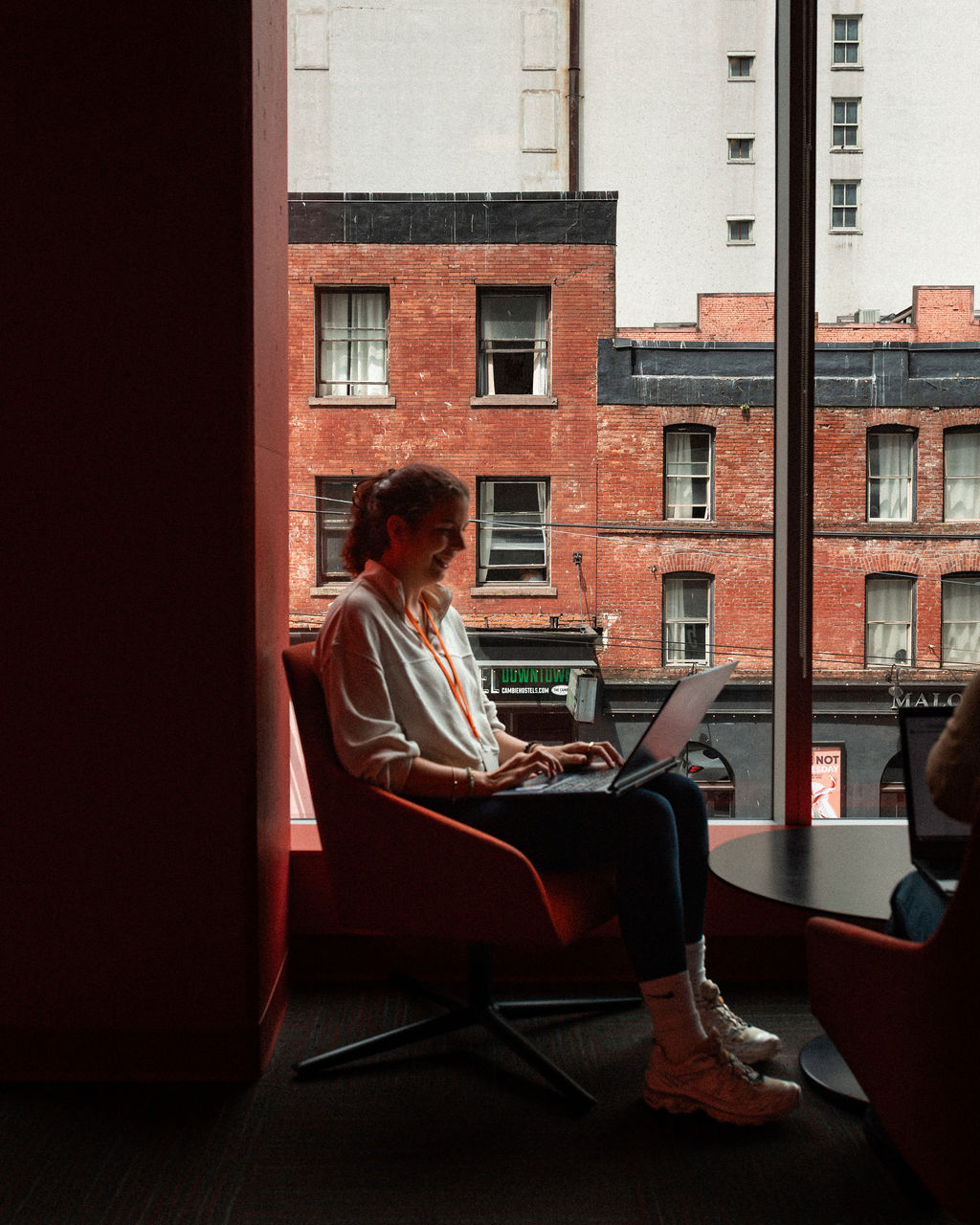 A person sits in a chair by a large window, working on a laptop, with brick buildings visible outside in daylight.