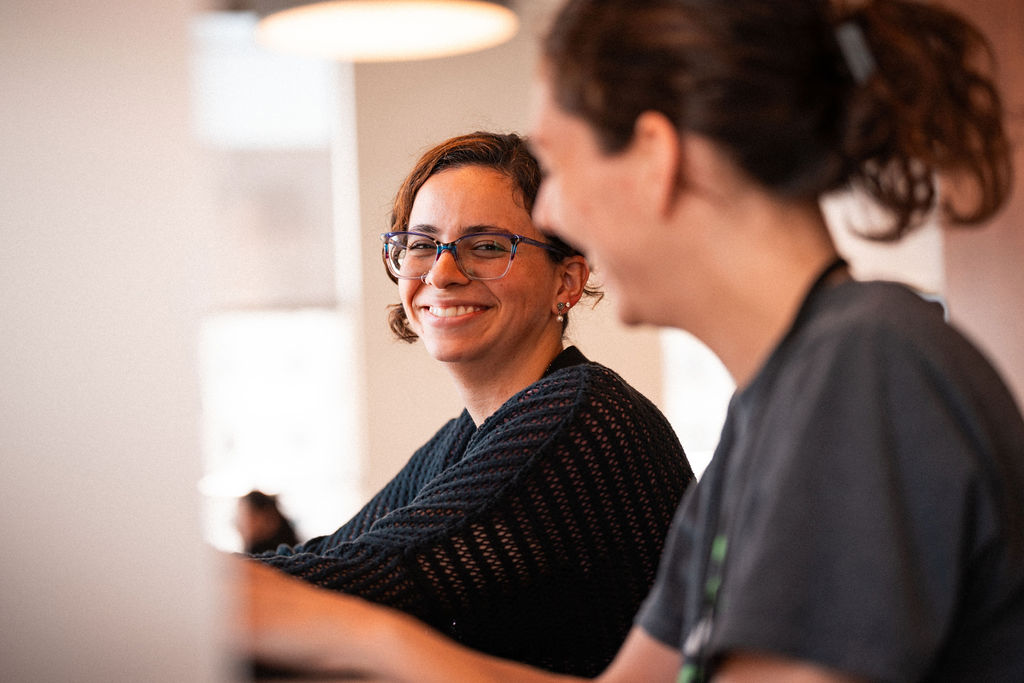 Two people sit indoors, one facing the camera and smiling while the other—perhaps an international student—is seen in profile, slightly out of focus.