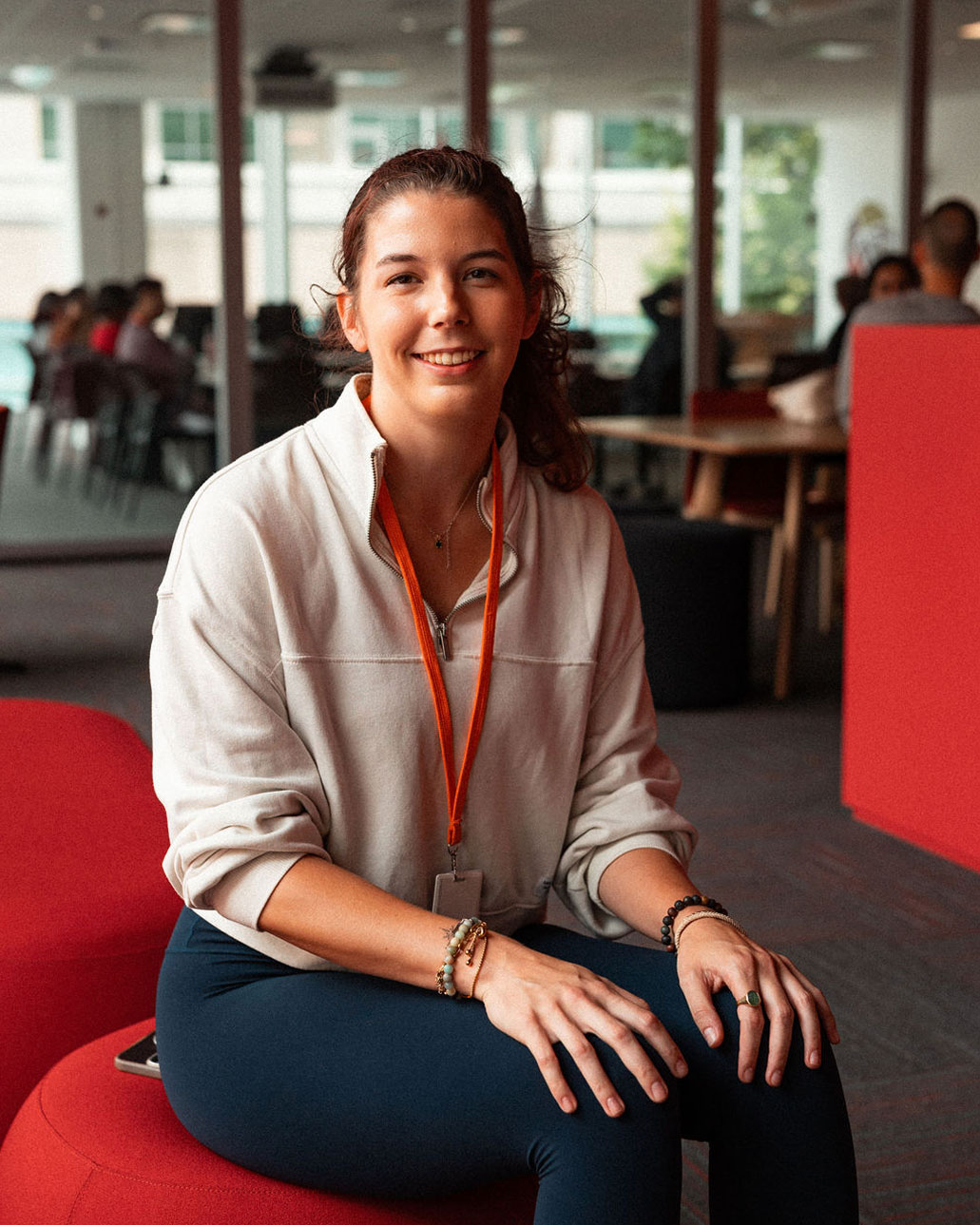 A woman with a lanyard sits on a red chair in a modern office space, smiling at the camera. The bright environment reflects the collaborative spirit of an Online Campus, with others engaging in the background.