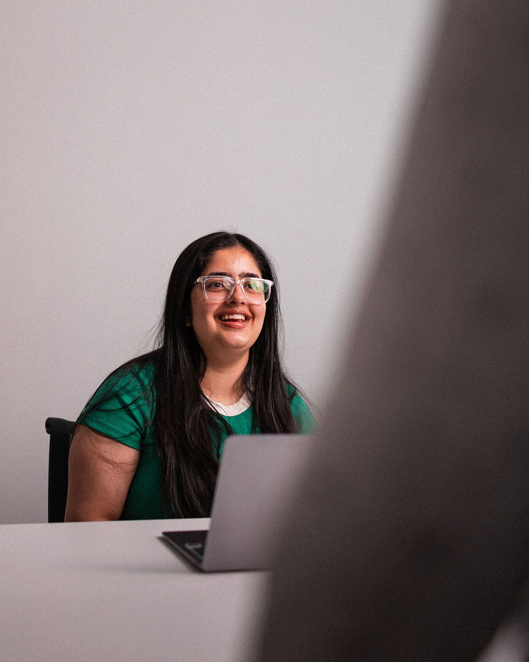 A woman with long dark hair and glasses sits at a desk with her laptop, smiling at someone standing in the foreground—capturing the welcoming spirit of the Online Campus.