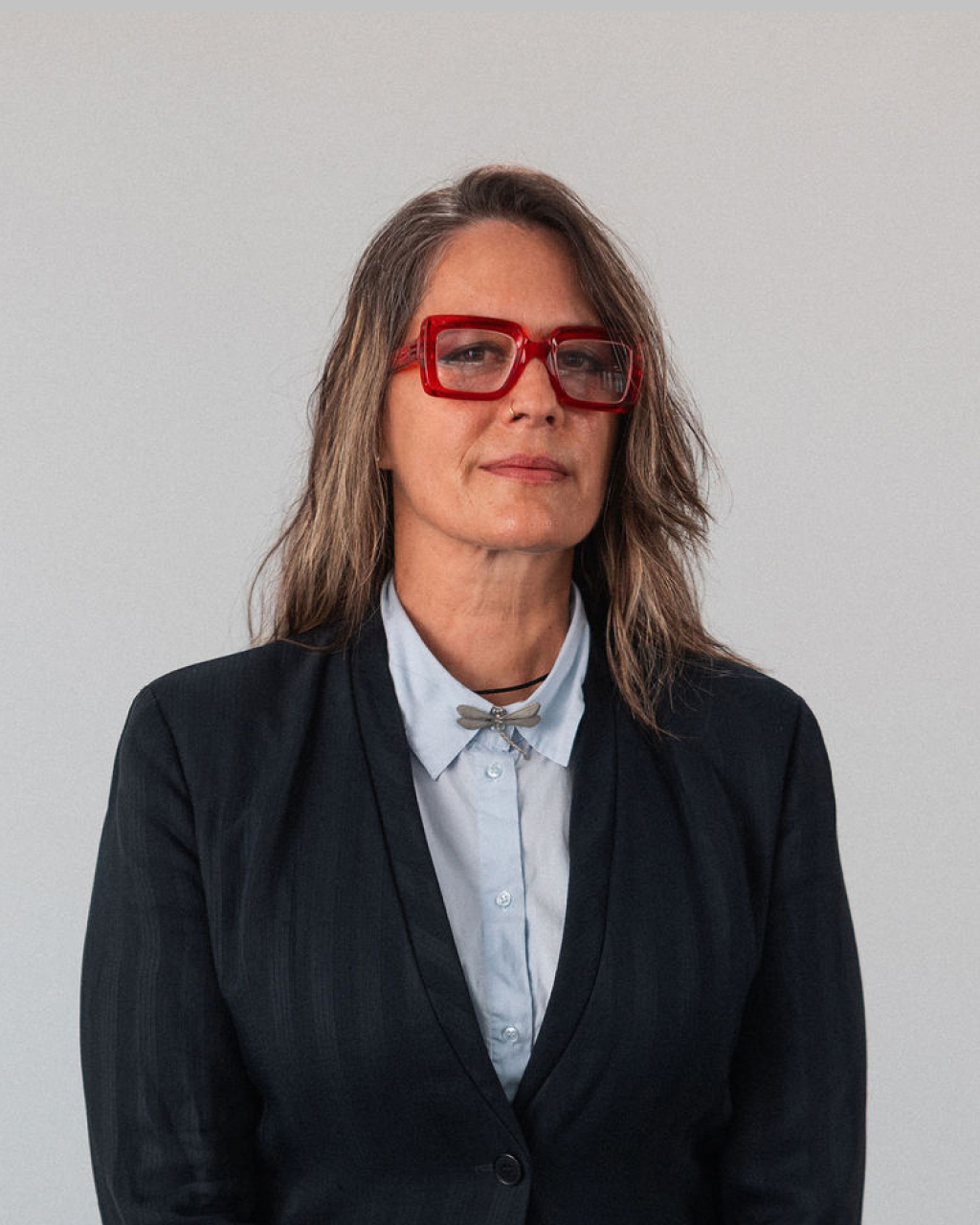 Asa-Sophia Maglio, a woman with long brown hair, red glasses, a black blazer, and a light blue shirt, stands against a plain light background.