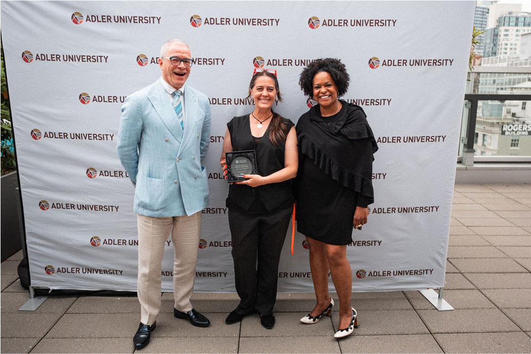 Three people, including the President, stand smiling in front of an Adler University backdrop; the person in the middle holds a plaque.