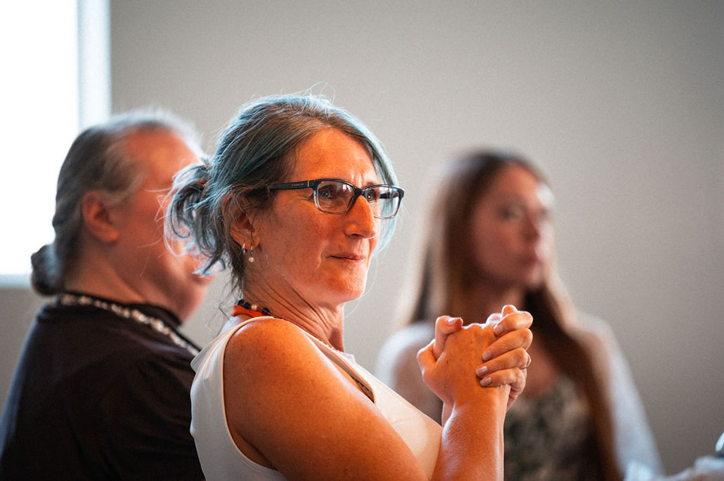 A woman with glasses sits at a table with two other people, hands clasped, looking forward in a brightly lit room, ready to discuss How To Apply for the upcoming opportunity.
