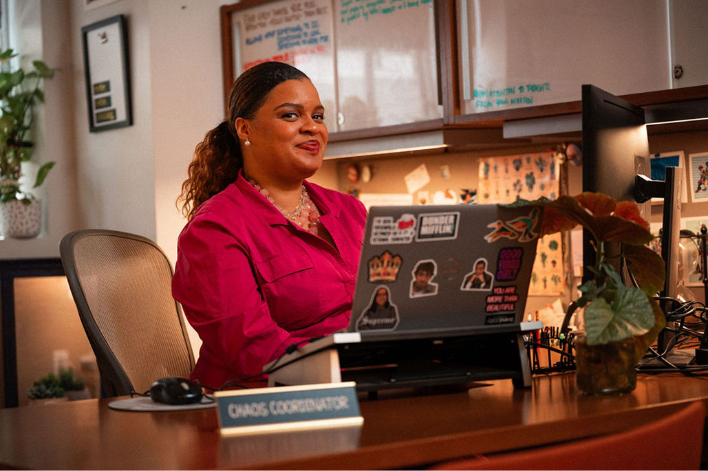 Woman in a pink shirt sits at a desk with a laptop covered in stickers, next to a nameplate reading "Chaos Coordinator" in a decorated office.
