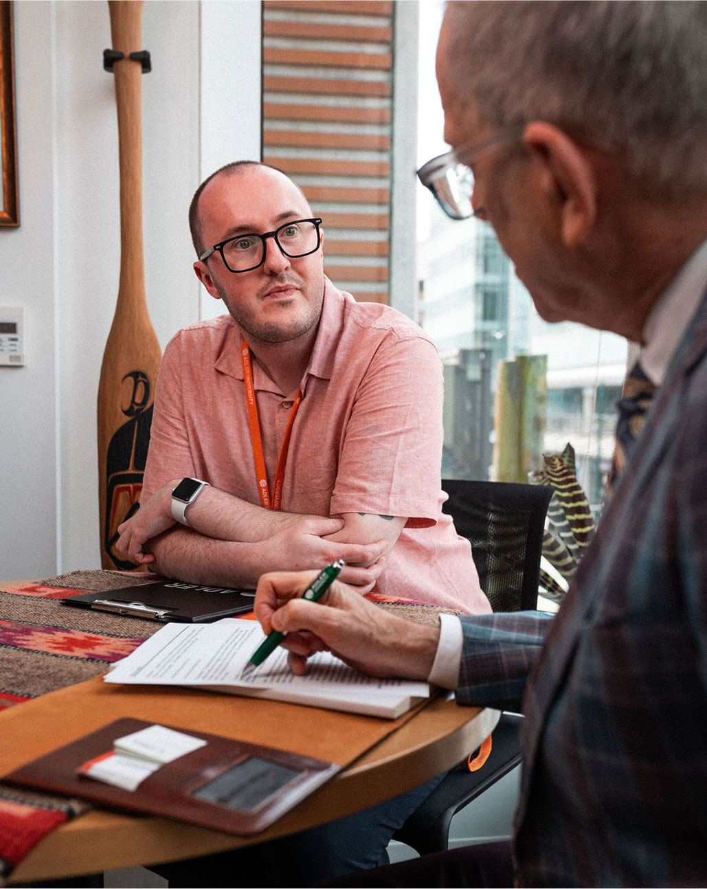 Two men sit at a table in an office; one in a pink shirt listens while the other, inspired by Alfred Adler's principles, writes thoughtfully in a notebook.