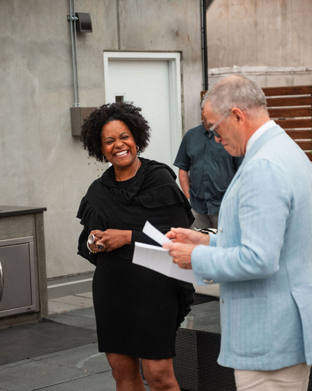 A woman in a black dress smiles while holding a cup, standing beside a man in a light blue jacket reading from papers. They are outdoors, near a staircase and building, possibly discussing ideas inspired by Alfred Adler.