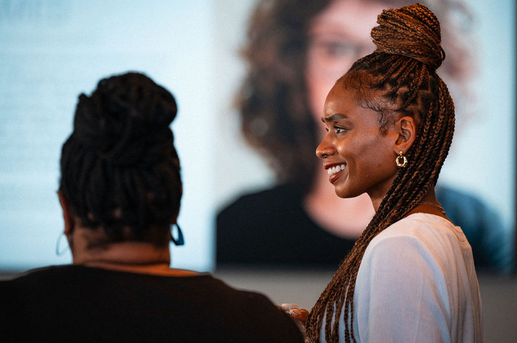 Two women with braided hair are seen from behind and in profile at an indoor event, with a blurred image of a person's face visible in the background.