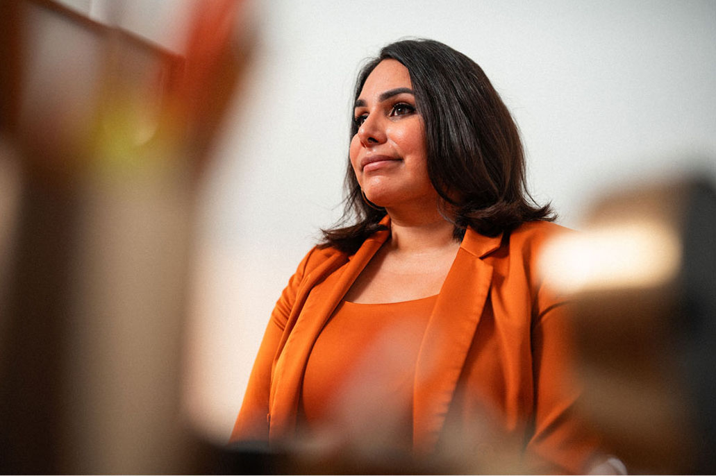 A woman with shoulder-length dark hair in an orange blazer and top looks thoughtfully to the side, embodying the drive and vision inspired by Faculty Research Funding, against a neutral background.