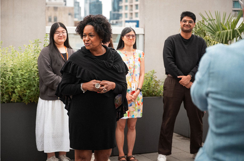 A woman in a black dress speaks outdoors to a small group; three people stand behind her, and one person is partially visible in the foreground.