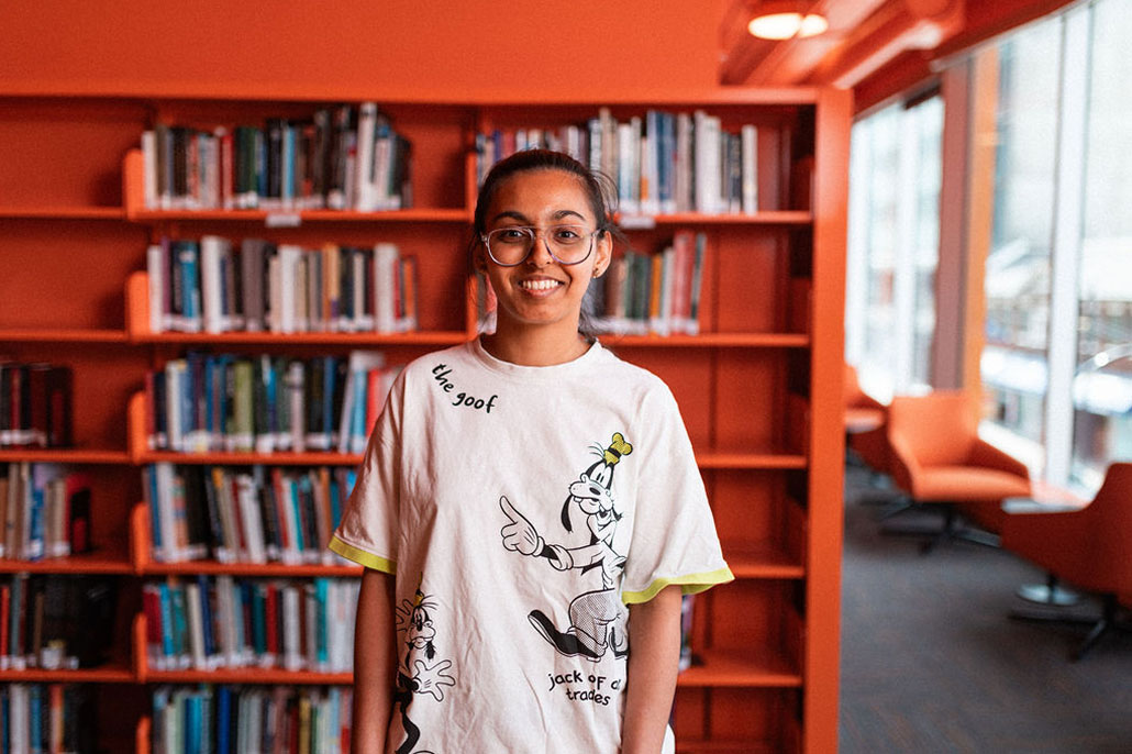 A person wearing a cartoon t-shirt and glasses stands in front of bookshelves in a brightly lit library with orange walls, embodying the spirit of giving opportunities through reading and learning.