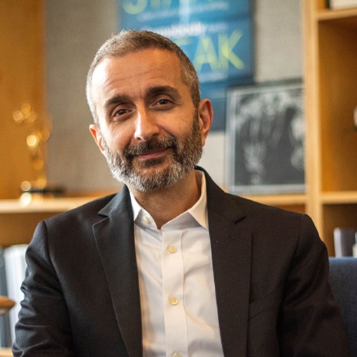 A man with short gray hair and a beard, wearing a white shirt and dark blazer, is seated in an office with shelves and framed items in the background.