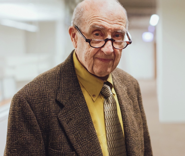 An elderly man wearing glasses, a brown blazer, yellow shirt, and patterned tie stands in a hallway, looking slightly to the side.