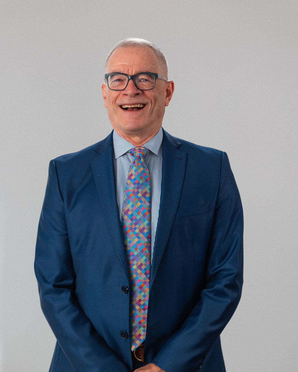 An older man with short gray hair and glasses is wearing a blue suit, striped shirt, and colorful patterned tie, standing and smiling against a plain light background.