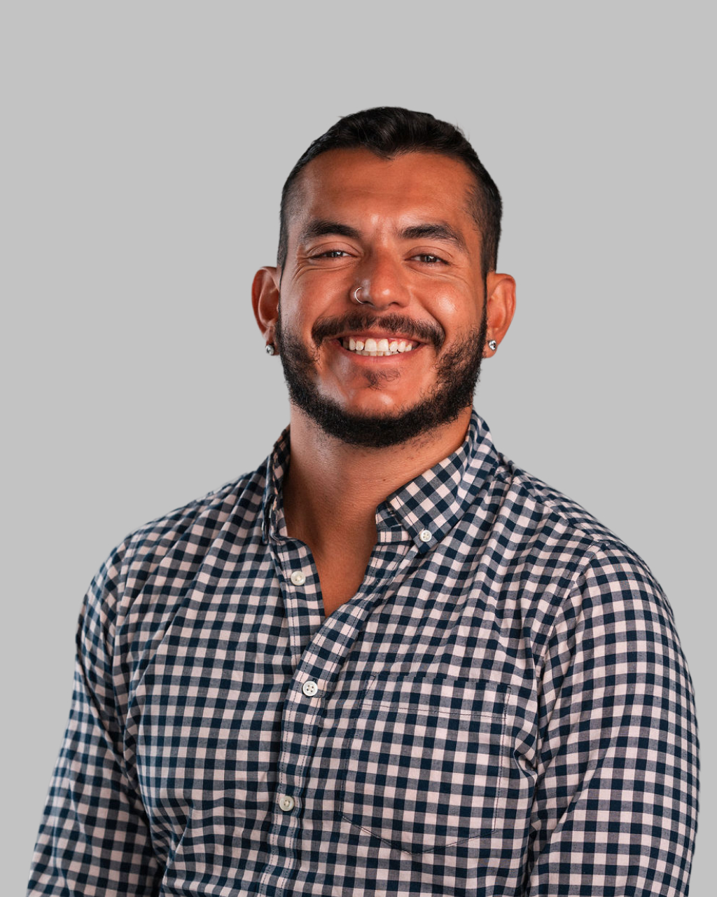 Cesar Perez, a man with short dark hair and beard, wears a blue and white checkered shirt and smiles at the camera against a plain grey background.