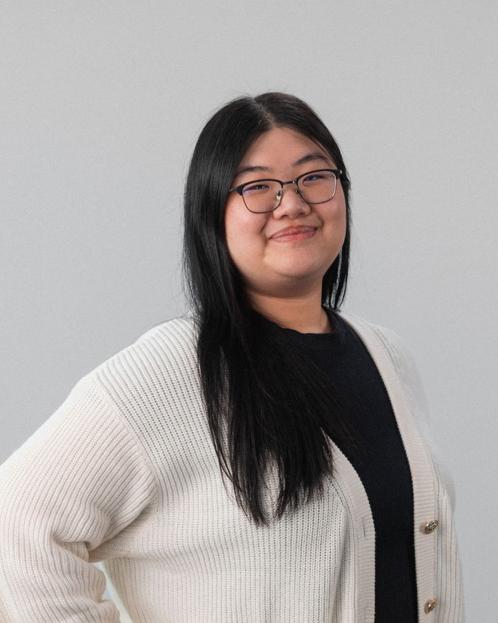 Charlene E., a person with long black hair and glasses, stands in front of a plain light background wearing a white cardigan and black top, smiling slightly.