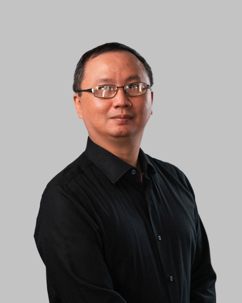 Chia-Chiang Wang, wearing glasses and a black button-up shirt, stands against a plain grey background, looking slightly to the side.