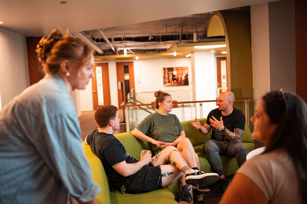 Five people sit and talk on green couches in a brightly lit, modern lounge area. Some are engaged in conversation while one person stands nearby, listening.