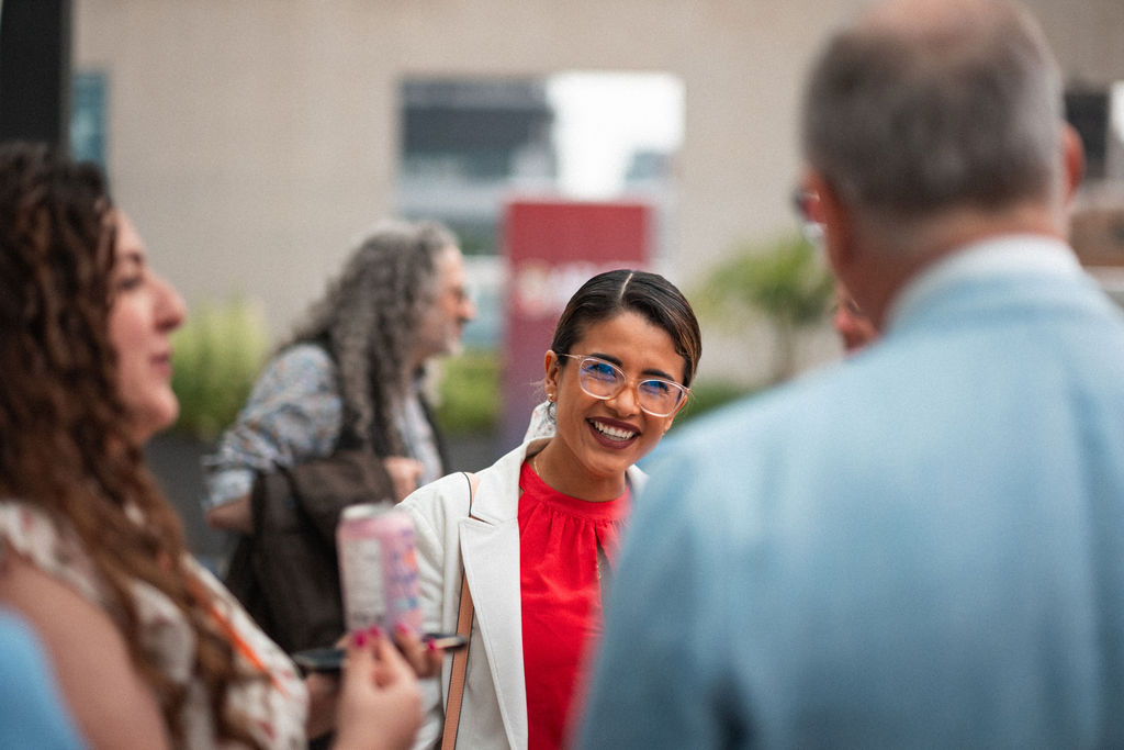 A woman wearing glasses and a red top smiles while conversing with people at an outdoor gathering.