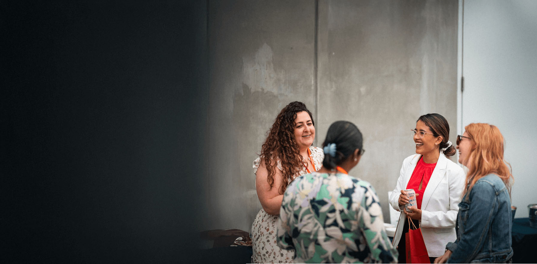 Four women stand in a group, talking and smiling, in front of a plain gray wall. The scene appears to be informal and social.